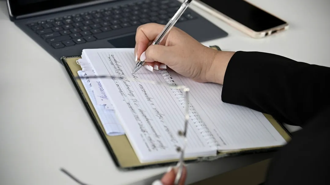 A person writing on a notebook with a laptop in the background