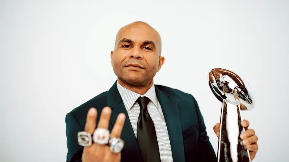 Confident man in a suit showcasing victory rings and trophy on a white background.