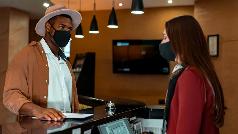 A man and woman in face masks checking in at a hotel reception, emphasizing safety protocols.